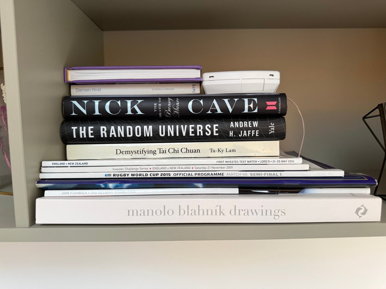 Bookshelf with several books and magazines laying horizontally, notably The Random Universe by Andrew H. Jaffe, as well as books by Nick Cave, Damian Hirst and Manolo Blahnik.